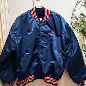 May include: A navy blue satin bomber jacket with red and white striped trim on the collar, cuffs, and hem. The name "Ray" is embroidered in red script on the chest. The jacket is hanging on a white hanger.