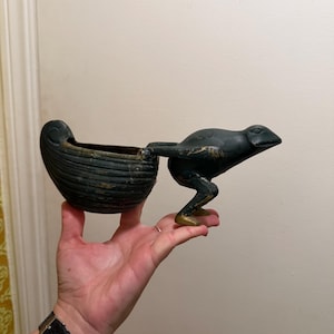 May include: A dark green, cast metal decorative bowl shaped like a small boat, attached to a stylized frog figure. The frog is positioned as if pushing the boat, with visible gold-toned wear on its feet and body, showing a weathered finish.