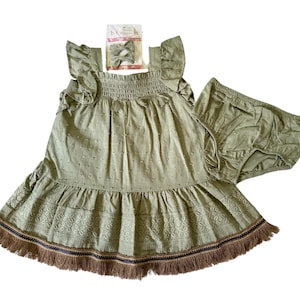 May include: A sage green baby dress set with matching bloomers and a bow. The dress features a ruffled neckline and hem with a floral embroidered design and a fringe trim. The bow is attached to a headband.