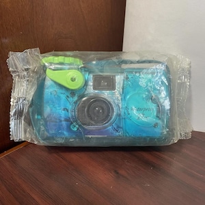 May include: A blue and teal waterproof film camera encased in clear plastic packaging. The camera features a bright green film advance knob and shutter button, a central lens, and the word Waterproof printed on the front casing in black text.