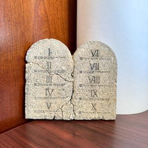 May include: Two stone tablets with the Ten Commandments inscribed in black text. The tablets are beige with a rough texture and a cracked appearance. Roman numerals and text are visible. The tablets are displayed on a dark wood surface.