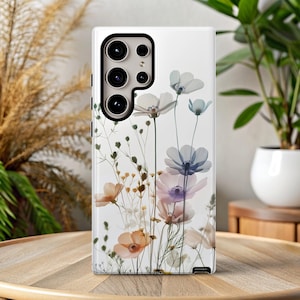 May include: A white phone case featuring a floral design with translucent flowers in shades of white, peach, purple, and blue. The case has a camera cutout and is set on a wooden table. The background includes plants and a neutral wall.
