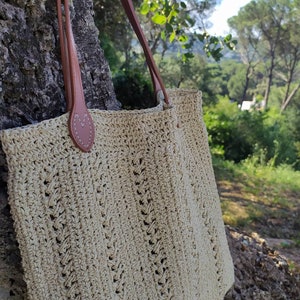 May include: A beige crocheted tote bag with brown leather straps. The bag has a simple, classic design and is perfect for carrying everyday essentials.