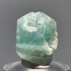 May include: A pale turquoise crystal with a hexagonal form. The surface of the crystal is rough and textured, with visible striations. The gemstone is displayed on a clear, round stand. The crystal has a slightly rounded top.