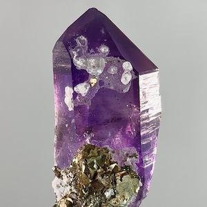 Puede incluir: Un vibrante cristal de amatista púrpura con una punta. El cristal tiene una base metálica dorada y plateada. Se ven inclusiones blancas y plateadas en la superficie. El cristal está sobre un fondo gris claro.