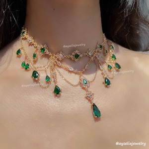 Könnte beinhalten: Goldfarbene Halskette mit smaragdgrünen Tropfen- und quadratischen Edelsteinen. Die Halskette hat filigrane Kettendetails und klare Kristalle. Der Text "@ayseliajewelry" ist sichtbar. Ein auffälliges Schmuckstück.