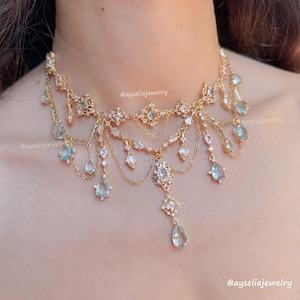 May include: A gold-toned necklace with multiple strands and dangling aquamarine-colored teardrop-shaped gems. The necklace features intricate filigree and small clear crystals. The text "@ayseliajewelry" is visible.