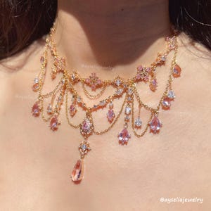 Puede incluir: Un collar dorado con piedras preciosas rosas y transparentes. El collar presenta múltiples capas de delicadas cadenas y gemas colgantes en forma de lágrima. El diseño incluye detalles florales y de estrella, creando un aspecto elegante y ornamentado.