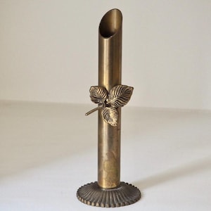Puede incluir: Un jarr&oacute;n de lat&oacute;n vintage con un adorno de hoja. El jarr&oacute;n tiene un cuerpo cil&iacute;ndrico con una parte superior acampanada y una base decorativa. El detalle de la hoja a&ntilde;ade un toque de elegancia inspirada en la naturaleza. El jarr&oacute;n mide unos 15 cm de alto.