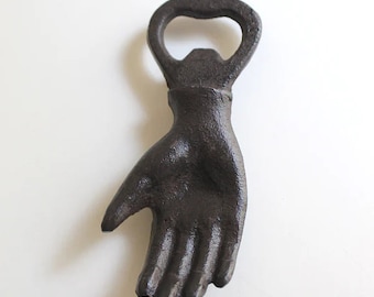 May include: A dark brown cast iron bottle opener shaped like a hand. The opener features a loop at the top for leverage and a detailed hand design. The product is designed to open bottles.