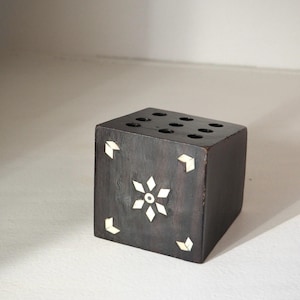 May include: A dark brown wooden cube with inlaid white accents. The cube features a floral design on one side and small geometric shapes on the corners. The top has seven circular holes.