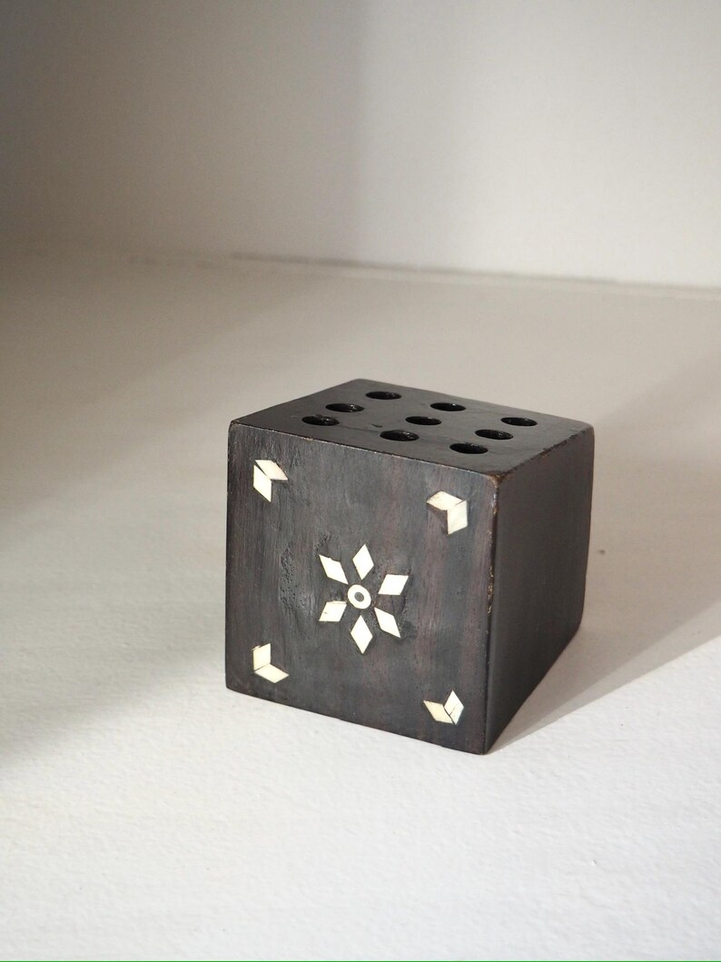 May include: A dark brown wooden cube with inlaid white accents. The cube features a floral design on one side and small geometric shapes on the corners. The top has seven circular holes.