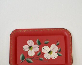 May include: A rectangular red metal tray with a hand-painted floral design. Two white dogwood flowers with green leaves and stems are centered on the tray. The tray has a scalloped gold border.