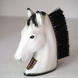 May include: A white ceramic horse head brush featuring black painted details on the ears and mane. The brush has stiff black bristles attached to the back of the horse head. A small, colorful crest emblem is visible on the lower neck area.