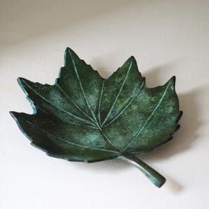 Puede incluir: Un plato decorativo en forma de hoja con una superficie texturizada y una pátina verde oscuro. El plato es de metal y tiene un tallo que se extiende desde la base. El diseño de la hoja está detallado con venas visibles.
