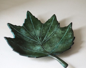 May include: A decorative leaf-shaped dish with a textured surface and a dark green patina. The dish is made of metal and has a stem extending from the base. The leaf design is detailed with visible veins.