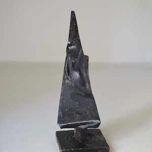 May include: A dark, weathered metal sculpture with a triangular shape, mounted on a square base. The sculpture has a rough texture and appears to be handmade. The base is also dark and square. The overall design is abstract.