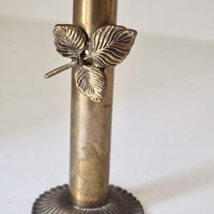 Puede incluir: Un candelero de lat&oacute;n vintage con un detalle de hoja. El candelero tiene una base redonda y acanalada y un cuerpo cil&iacute;ndrico. Un dise&ntilde;o de hoja de lat&oacute;n adorna el tallo, agregando un toque decorativo.