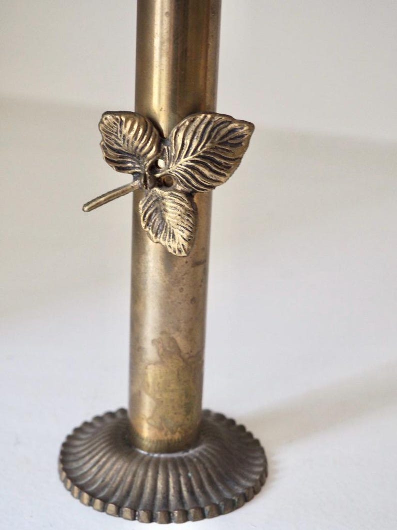 Puede incluir: Un candelero de lat&oacute;n vintage con un detalle de hoja. El candelero tiene una base redonda y acanalada y un cuerpo cil&iacute;ndrico. Un dise&ntilde;o de hoja de lat&oacute;n adorna el tallo, agregando un toque decorativo.