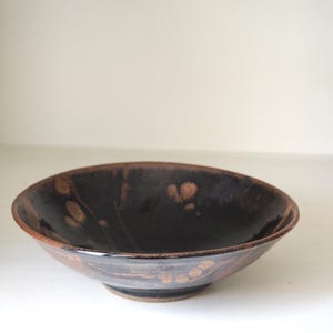 May include: A ceramic bowl with a dark brown interior and exterior, featuring a glossy finish. The bowl has a wide, shallow shape with a slightly raised base and a rim with a lighter brown color. The bowl has a decorative pattern.