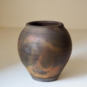 Op de afbeelding: Een handgemaakte, ronde keramische vaas met een brede opening. De vaas heeft een donkerbruine buitenkant met hints van goud en oranje, wat een rustieke en aardse uitstraling creëert. De vaas is geschikt voor het tentoonstellen van bloemen of als decoratief stuk.