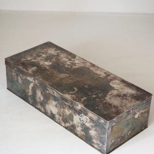 Könnte beinhalten: Eine rechteckige Aufbewahrungsbox aus Metall mit einer verwitterten, angelaufenen Oberfläche. Die Außenseite zeigt ein fleckiges Muster aus Dunkelgrau, Silber und oxidierten Blau-Grün-Tönen, was auf ein hohes Alter und Abnutzung hindeutet.