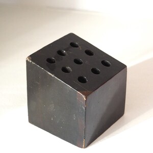 May include: A dark brown wooden cube with nine evenly spaced holes on the top surface. The cube has a slightly worn appearance, with visible wear on the edges. The object is likely a desk organizer or pen holder.
