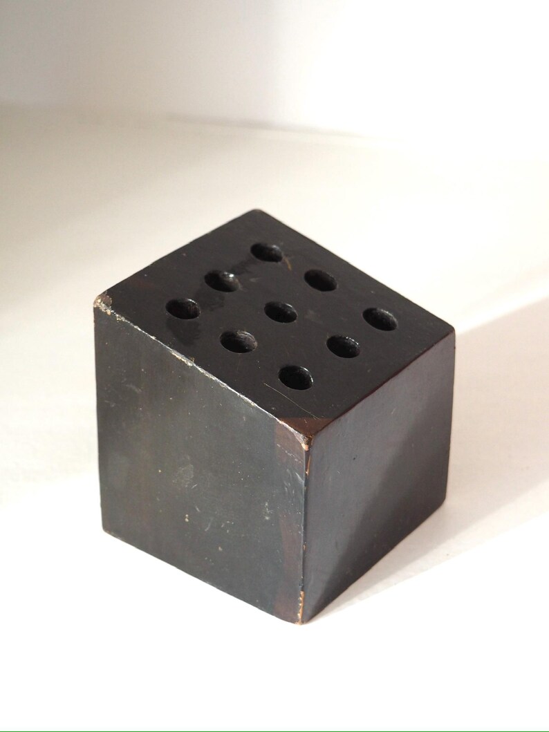 May include: A dark brown wooden cube with nine evenly spaced holes on the top surface. The cube has a slightly worn appearance, with visible wear on the edges. The object is likely a desk organizer or pen holder.