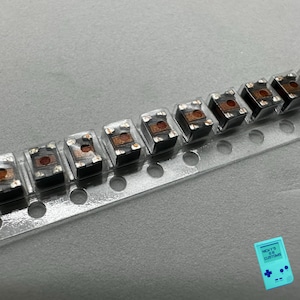 May include: A close-up of a clear plastic strip with ten small, square, black electronic components. Each component has a copper-colored center and is arranged in a row.