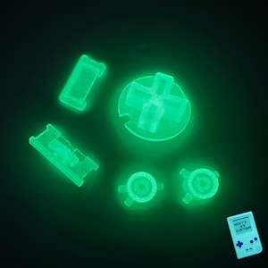 May include: A set of glow-in-the-dark green buttons for a game console. The buttons are shaped like a cross, a circle, and a rectangle. The buttons are on a black background. The text on the game console says "NICKY'S 8 BIT CUSTOMS".