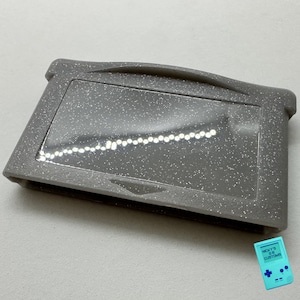 May include: A gray Game Boy Advance game cartridge with a glittery finish. The cartridge has a clear plastic window that shows the game's internal components.