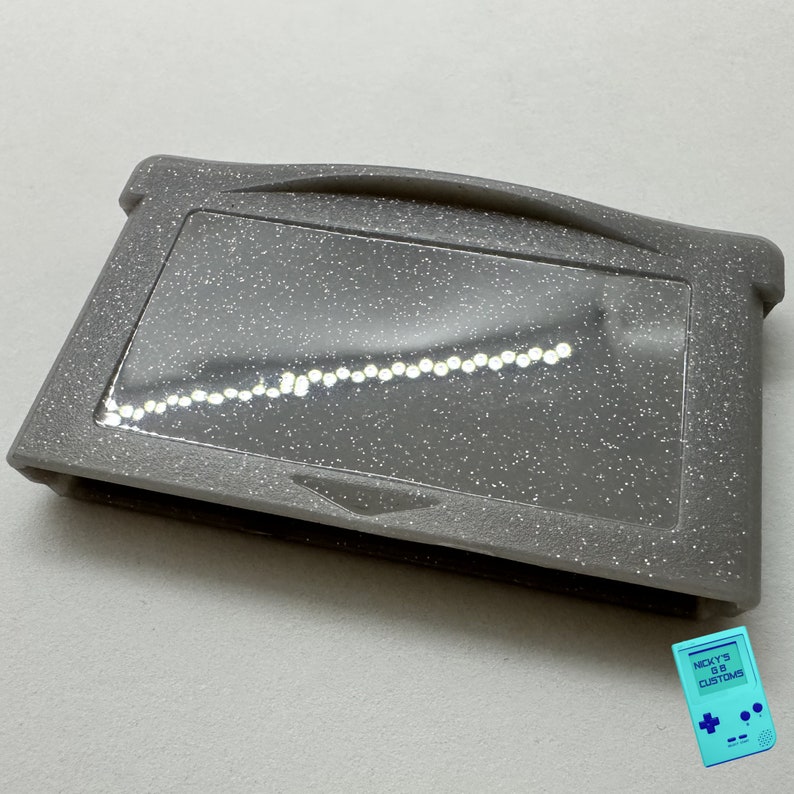 May include: A gray Game Boy Advance game cartridge with a glittery finish. The cartridge has a clear plastic window that shows the game's internal components.