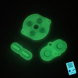 May include: Four glow-in-the-dark green replacement buttons for a video game controller. The buttons are shaped like circles, squares, and an 8-way directional pad. The buttons are on a black background.
