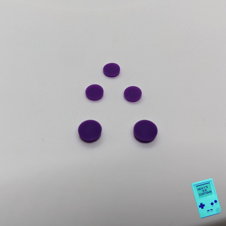Peut inclure: Cinq boutons ronds violets dispos&eacute;s en forme de triangle sur une surface blanche.
