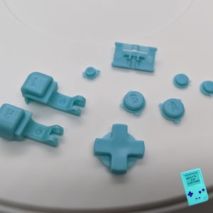 May include: A set of light blue replacement buttons for a game console controller. The set includes a directional pad, four face buttons, two shoulder buttons, and a start button. The buttons are made of plastic and have a smooth, matte finish.