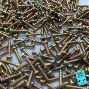 May include: A large pile of gold-colored screws, scattered on a white surface. The screws are all the same size and shape, and they are arranged in a random pattern.