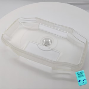 May include: A clear plastic case for a handheld gaming device. The case has a raised area for the device's screen and buttons. The case is designed to protect the device from scratches and bumps.