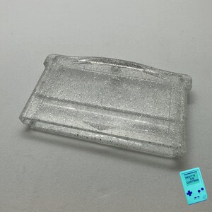 May include: A clear plastic Game Boy Color game cartridge case with a silver glitter finish.
