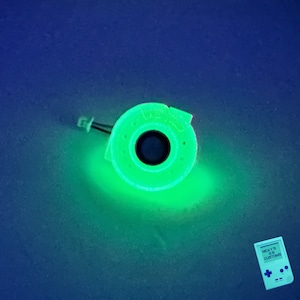 May include: A green glow-in-the-dark plastic brake rotor keychain with a black center. The keychain is on a blue surface.