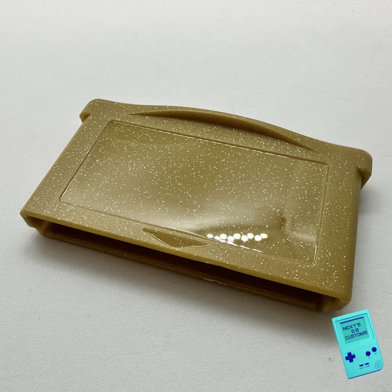 May include: A gold Game Boy Advance game cartridge case with a glittery finish.