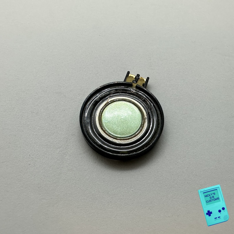 May include: A black round speaker with a green center. The speaker has a gold colored wire attached to it.