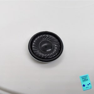 May include: A black round speaker with a silver rim. The speaker has a black center and a series of concentric circles radiating outward.