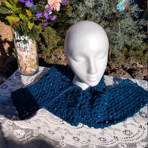 May include: Teal blue crocheted infinity scarf with a sparkly texture. The scarf is displayed on a white lace tablecloth.
