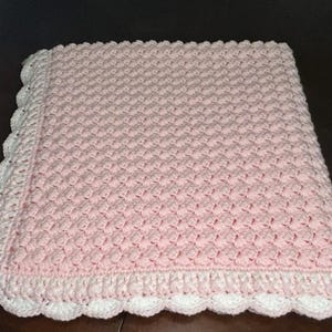 May include: A pink crocheted baby blanket with a white crocheted border. The blanket has a textured, diamond-shaped pattern.