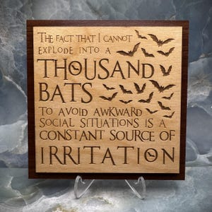 May include: Wooden sign with a dark brown frame and light brown center. The sign reads "The fact that I cannot explode into a thousand bats to avoid awkward social situations is a constant source of irritation."
