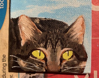 May include: A close-up painting of a tabby cat's face with green eyes. The cat is looking directly at the viewer. The background is a light blue sky with white clouds.