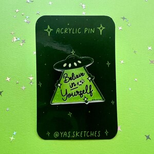 May include: Green and black acrylic pin with a UFO and a triangle with the text "Believe in Yourself". The pin is on a black background with white stars. The text "Acrylic Pin" and "@YAS.SKETCHES" are also on the background.