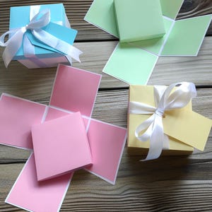 May include: An overhead shot showcases several colorful gift boxes. A light blue box with a white ribbon and bow sits near a pink box with open flaps. A yellow box with a white ribbon and tag is also visible, along with a green box.