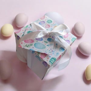 May include: A square gift box with a white ribbon tied in a bow. The box is decorated with a pattern of pastel Easter eggs and floral accents. Several pastel-colored eggs are scattered around the box on a pink background.