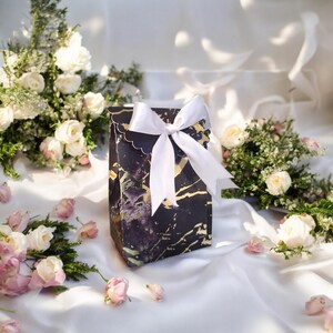 May include: A gift bag with a black marble design and gold accents, tied with a white satin bow. The bag has a scalloped edge and is surrounded by white and pink roses on a white satin surface.